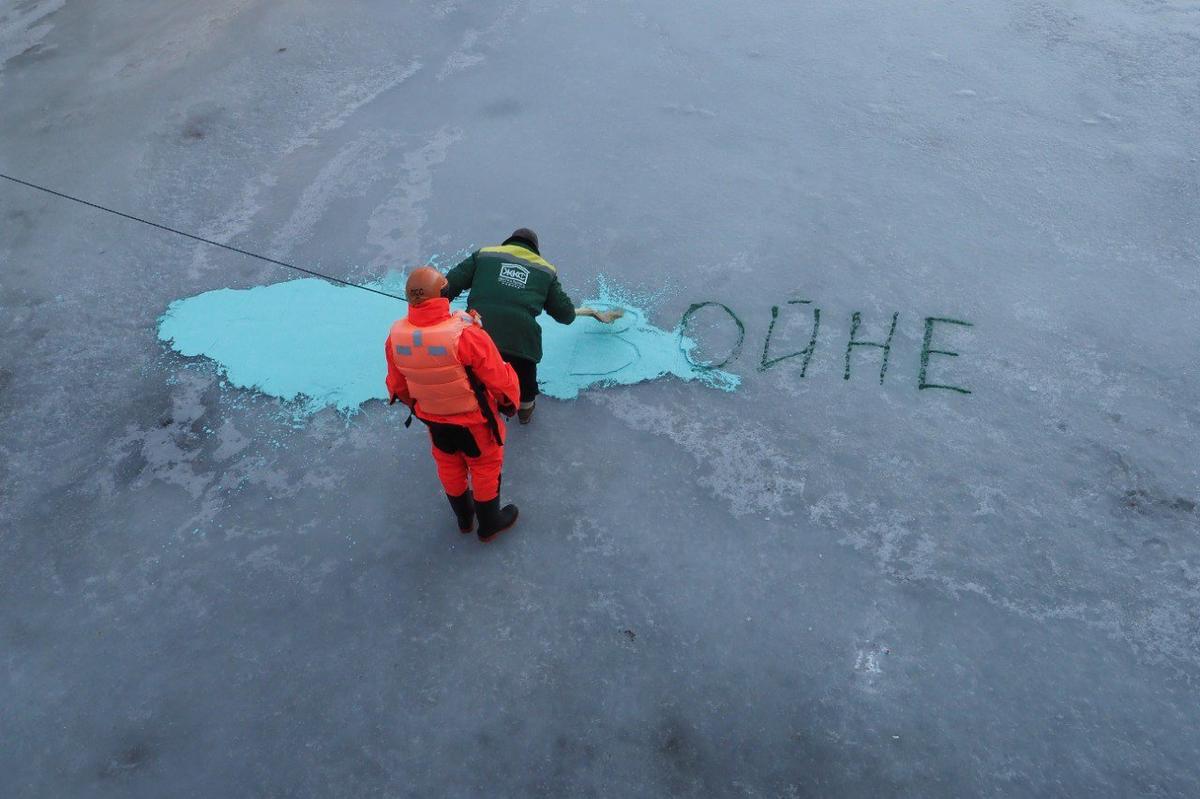 Коммунальщик под присмотром спасателя веником закрашивает зелёной краской надпись на льду Мойки. 6 марта. Фото: Олег Золото / MR7